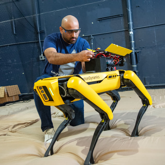 A Nevada engineering student wears protective eyewear and kneels while working on a yellow Boston Dynamics robot.